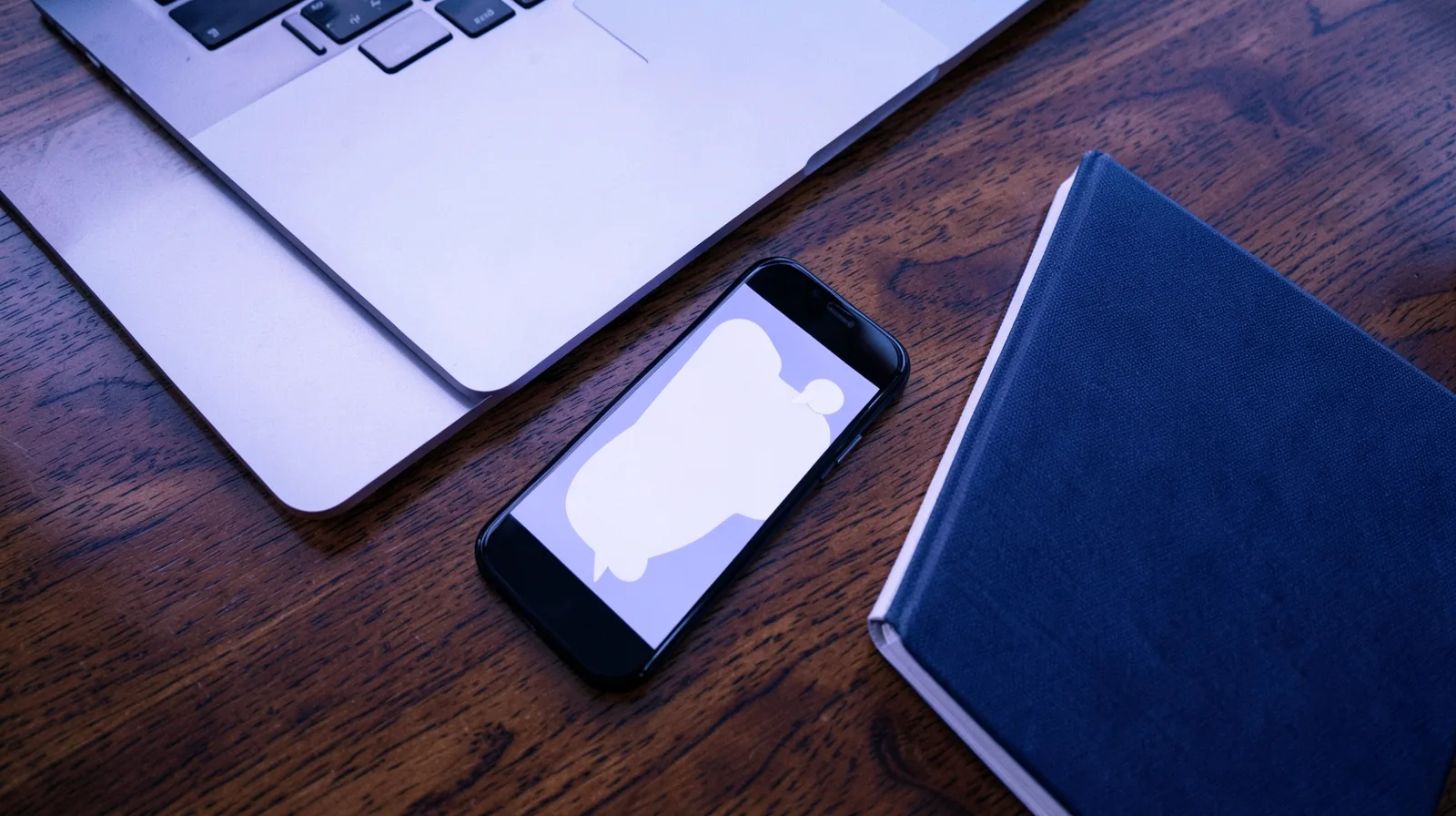 Celular com um balão de conversa vazio na tela, ao lado de um notebook e de um caderno fechado sobre uma mesa de madeira