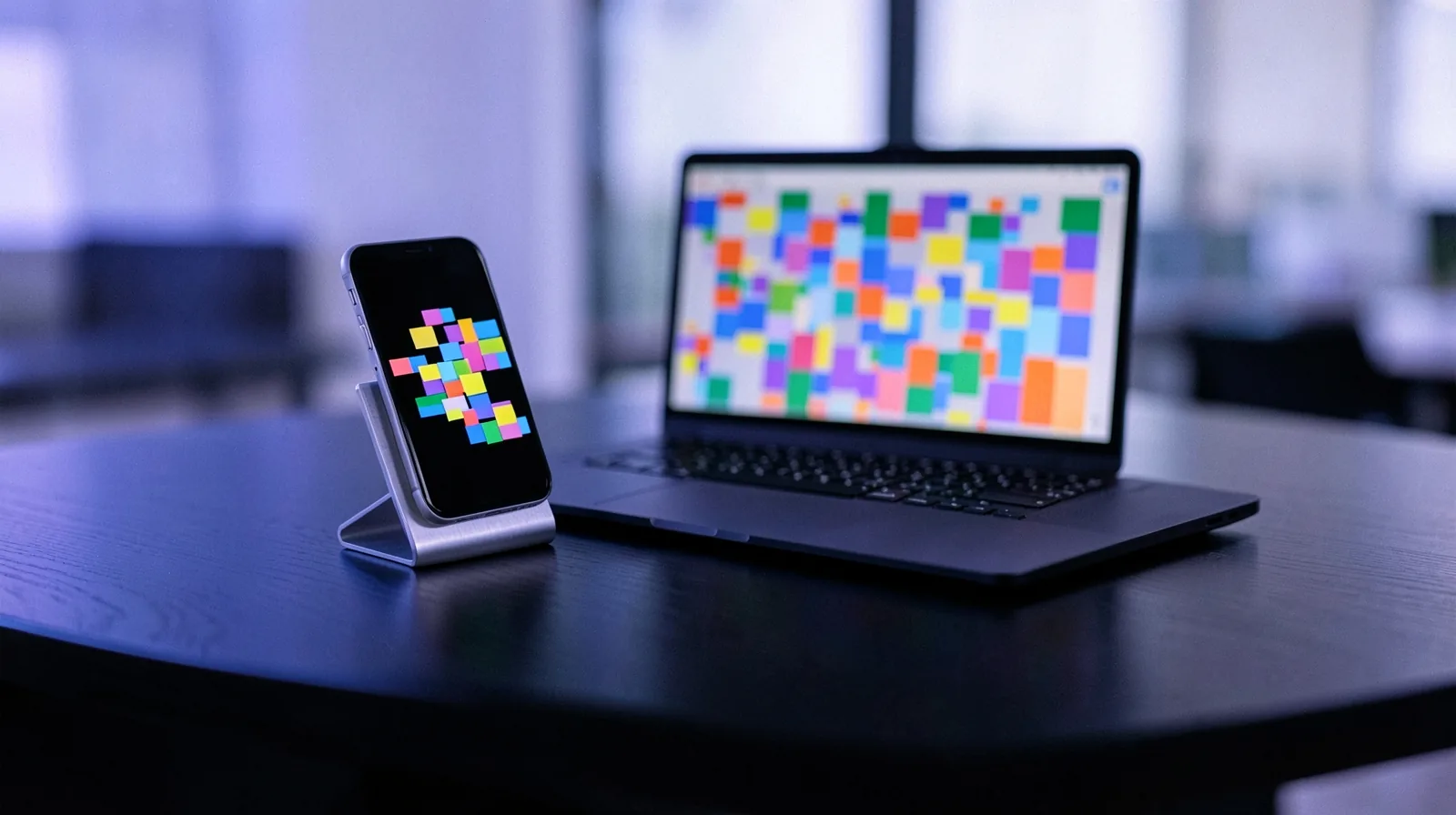 Celular em um suporte com uma grade de publicações em blocos coloridos, ao lado de um notebook com uma grade de blocos coloridos sobre uma mesa de escritório